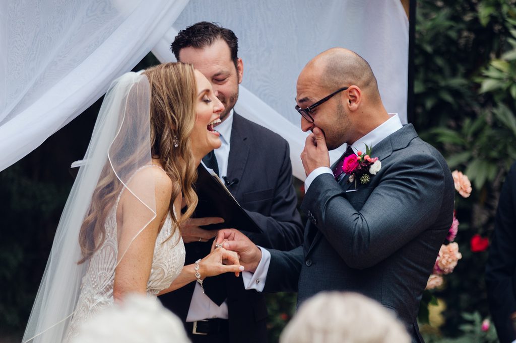 Bride and groom in a candid photo during their wedding ceremony. The bride is laughing, while the groom is emotional while putting her ring on her finger.