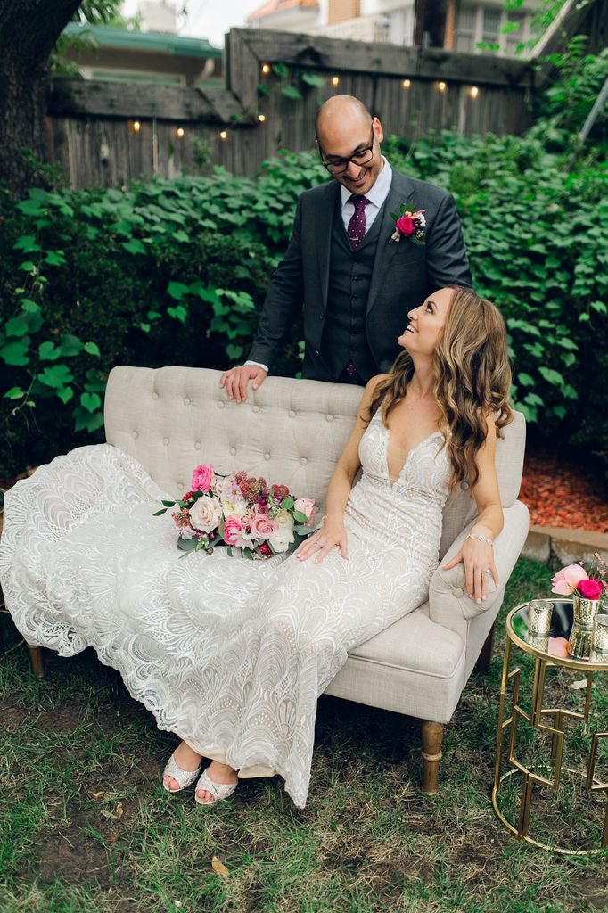 A bride is sitting on a settee with the groom standing behind her. The bride and groom are smiling naturally looking at each other.
