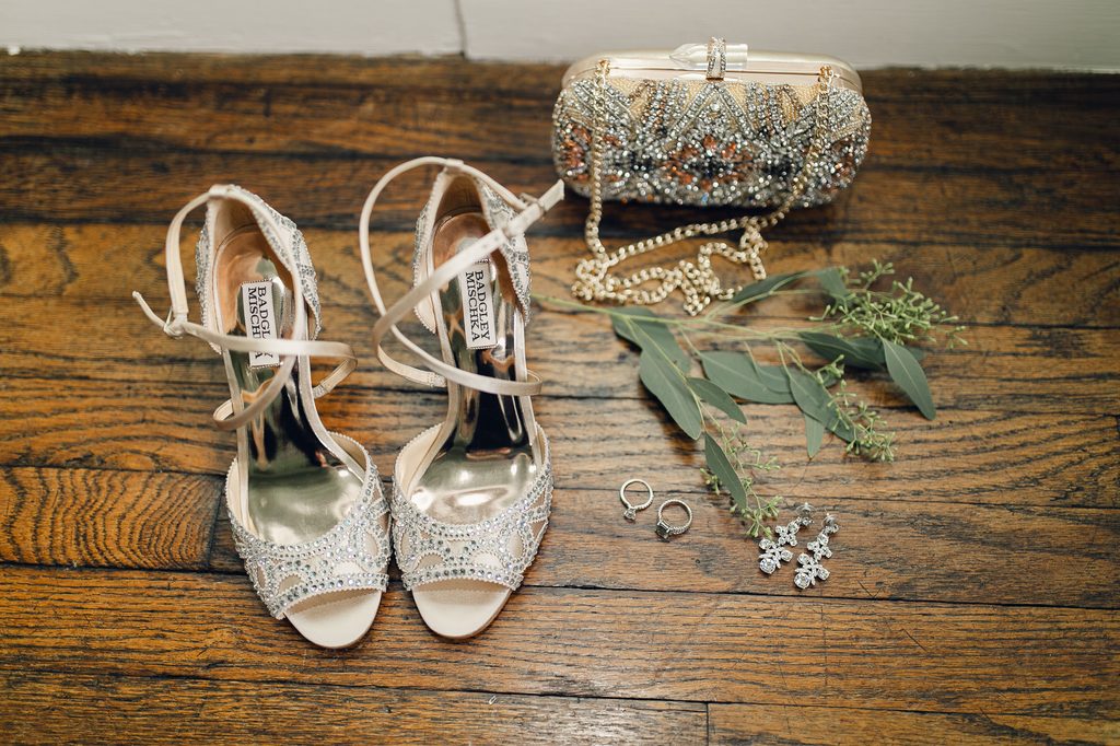 Embellished Badgley Mischka bridal heels, a beaded clutch, rings, and earrings arranged on a wooden floor. 