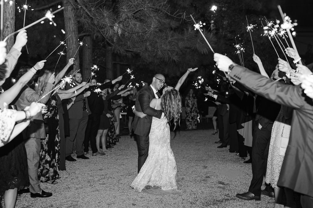 Black-and-white photograph of Megan and Keivan embracing between guests holding sparklers at their nighttime send-off from Dresser Mansion