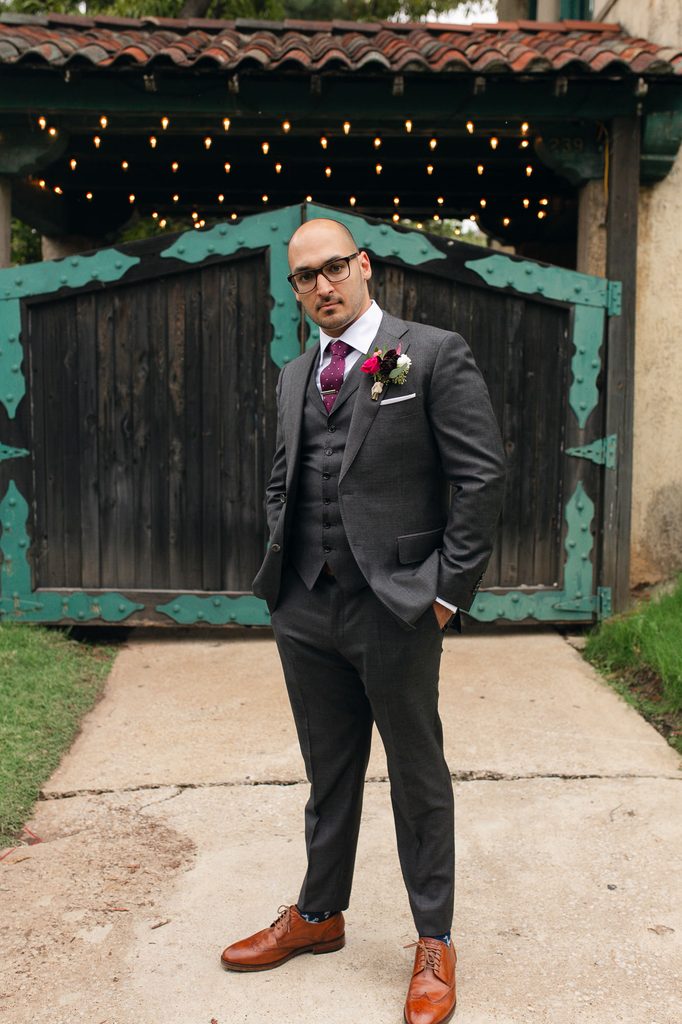 Keivan in a gray three-piece suit standing before wooden courtyard gates with blue-green trim. 
