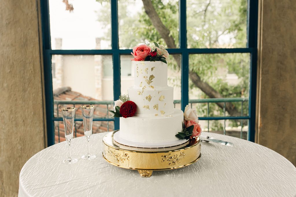 Three-tier white wedding cake with gold accents and pink and burgundy flowers on an ornate gold stand. 
