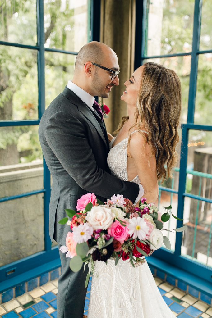 Megan and Keivan smiling at each other beside blue-green windows, with her colorful bouquet in the foreground for their wedding at Dresser Mansion.