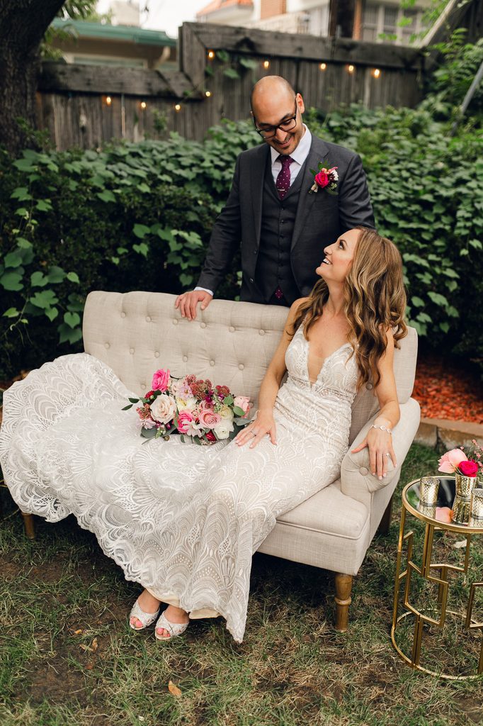 Megan seated in a garden chair with her lace train spread out and Keivan standing beside her. 