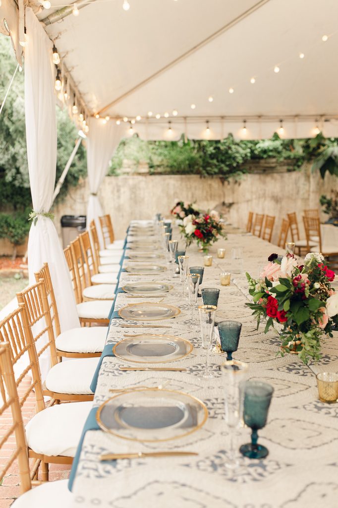 Reception table with patterned linens, blue goblets, gold-rimmed plates, and flowers beneath a canopy with string lights. 