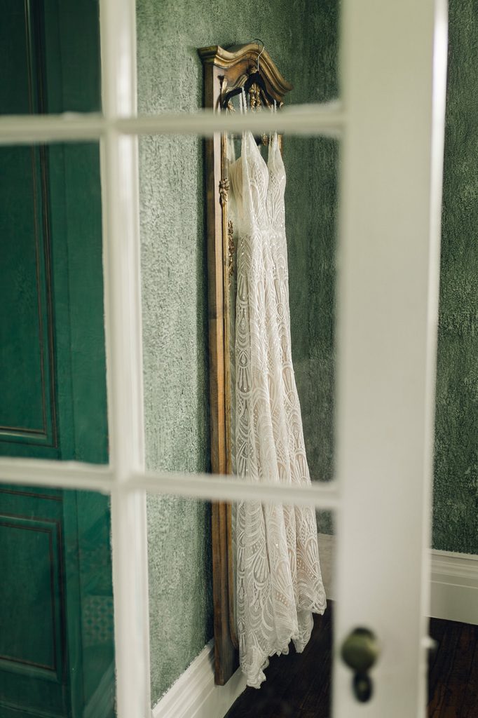 Lace wedding gown hanging in a green room, photographed through a glass-paneled door. 