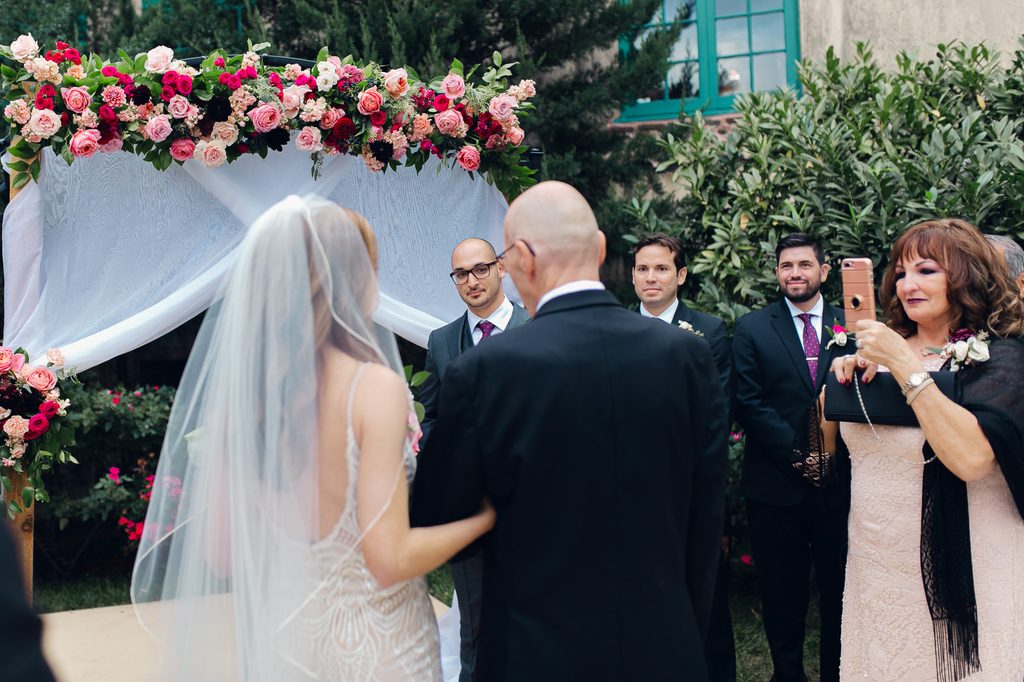 Keivan watching Megan approach with her father beneath a backdrop of pink flowers and white draping. 