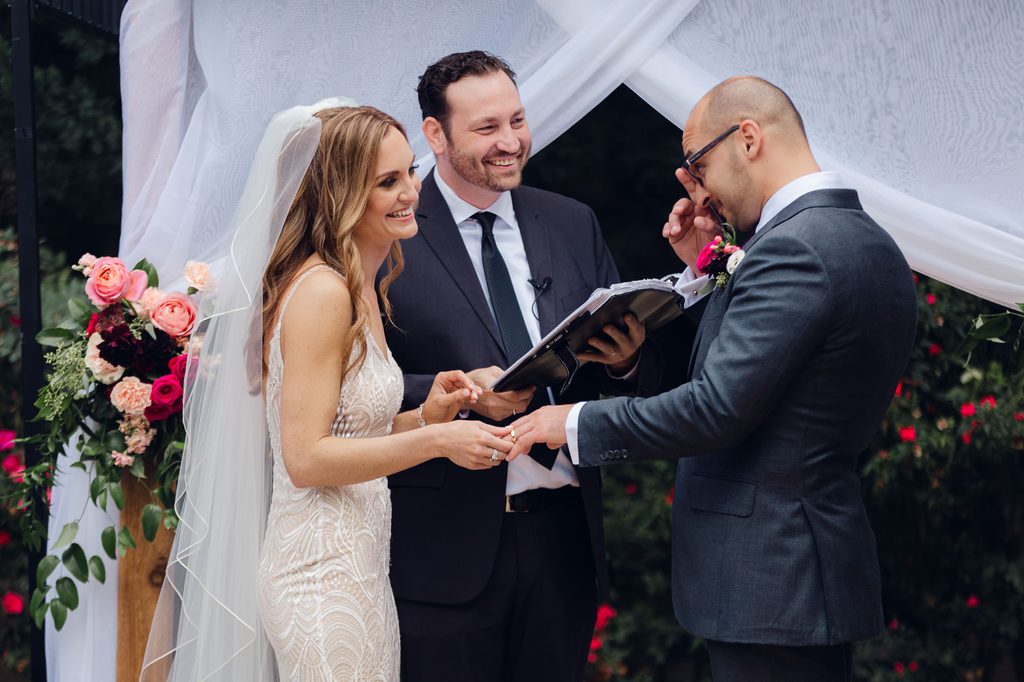 Megan smiling and holding Keivan’s hand during the ring exchange as he brings his other hand to his face. 
