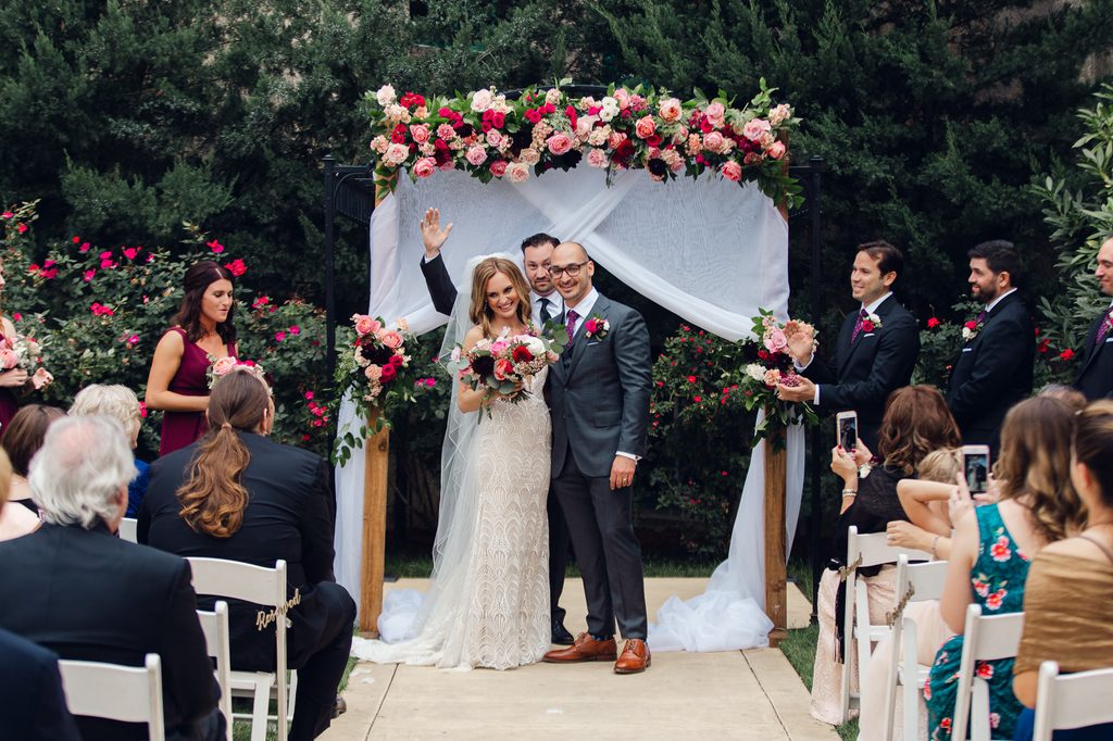 Megan and Keivan standing together beneath the floral ceremony backdrop as their wedding party applauds. 