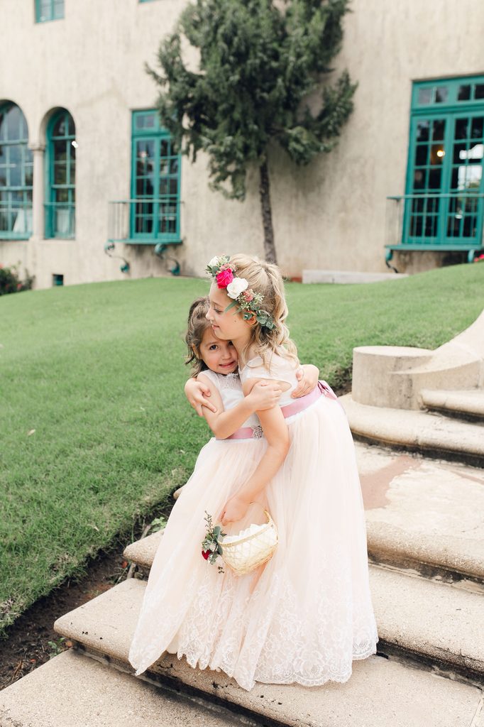 Two flower girls hugging on the mansion steps in pale dresses with pink sashes. 