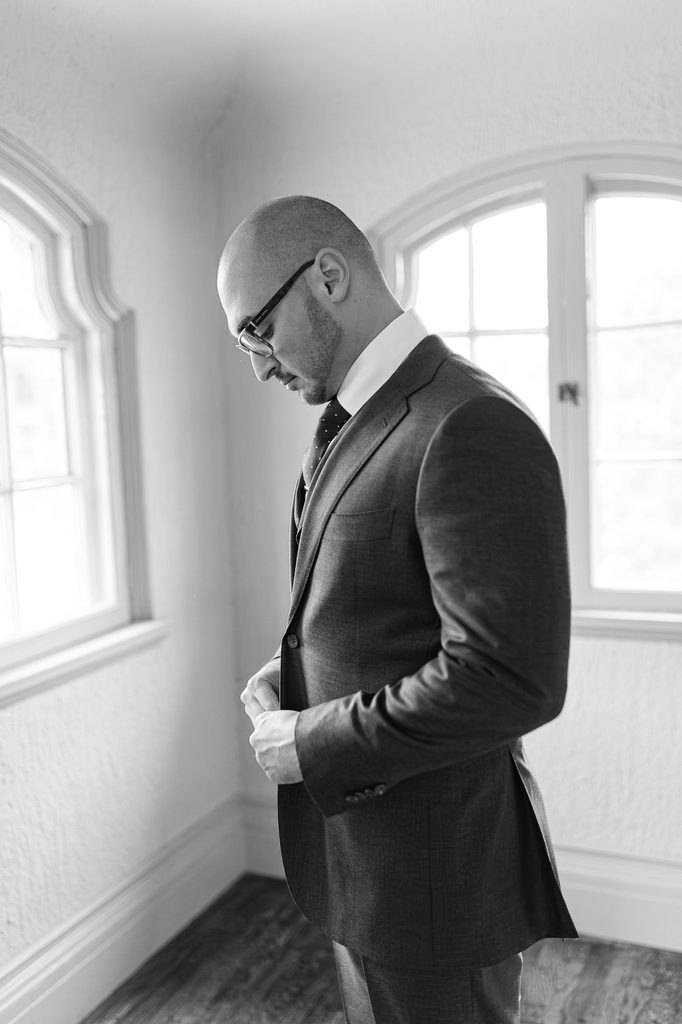 Black-and-white portrait of Keivan fastening his suit jacket beside arched windows. 