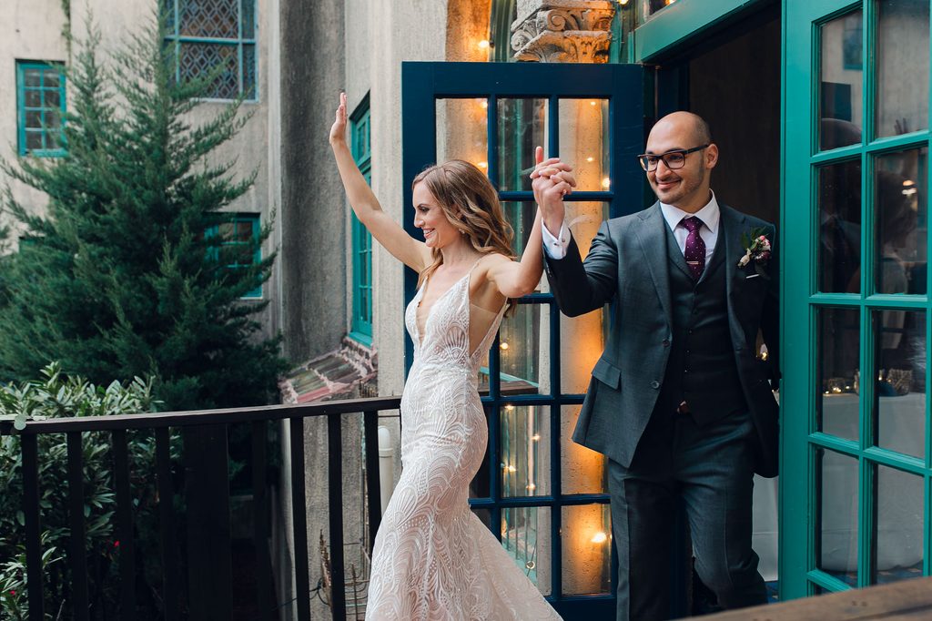 Megan and Keivan raising their joined hands as they step through the mansion’s blue-green doors. 